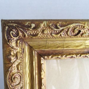 May include: Close-up of a corner of a vintage gold picture frame. The frame has ornate, raised detailing with a distressed finish, showing signs of age and wear. The frame's color is a mix of gold and brown.