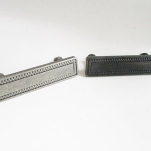 May include: Two cabinet pulls with a rectangular shape and a braided border. The pull on the left is silver and the pull on the right is black.