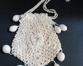 May include: Cream-colored crocheted drawstring bag with a floral design in the center. The bag is adorned with small, teardrop-shaped embellishments and a long, braided strap. The bag is likely handmade and has a vintage aesthetic.