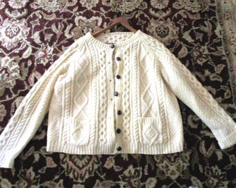 May include: An off-white cable knit cardigan sweater featuring a round neckline, long sleeves, and a front button closure with dark brown buttons. The garment includes two front patch pockets and a textured pattern of vertical cables and diamonds.