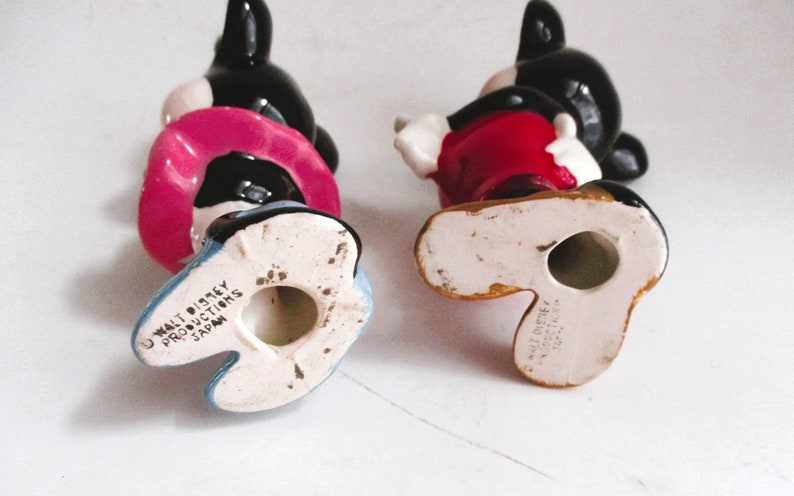 May include: Two ceramic Mickey Mouse figurines, one in red and one in pink, with black ears and shoes. The bottom of each figurine has the text "Walt Disney Productions Japan".