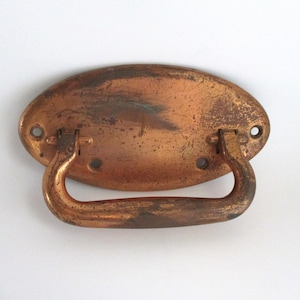 May include: A vintage-style copper drawer pull with an oval backplate and a curved handle. The copper has a distressed finish with visible wear and patina, showing shades of brown and green. The pull has four screw holes for mounting.