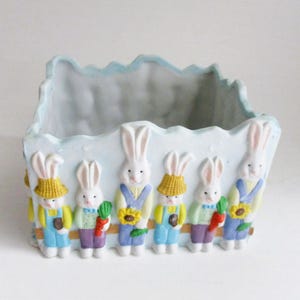 May include: A rectangular ceramic planter with a light blue exterior and a scalloped top edge. The planter is decorated with six white bunny figures wearing overalls and holding carrots and sunflowers. The bunnies are painted in bright colors.