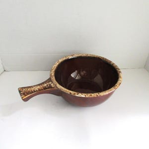 May include: A brown ceramic soup bowl with a handle. The bowl has a dark brown interior and a lighter brown rim. The handle is also brown with a textured design. This bowl is perfect for serving soups and stews.