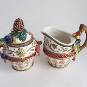 May include: A white ceramic sugar and creamer set with a winter holiday theme. The creamer features a twig handle with a red and yellow bird perched on it. The sugar bowl has a pine cone lid with green leaves and red berries.