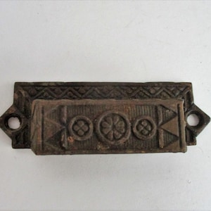 May include: A vintage cast iron drawer pull with a decorative design. The pull is brown and has a geometric pattern.