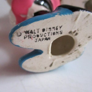 May include: Close-up of the bottom of a ceramic figurine. The figurine is painted blue, black, and pink. The bottom of the figurine is white with black writing that reads "Walt Disney Productions Japan".
