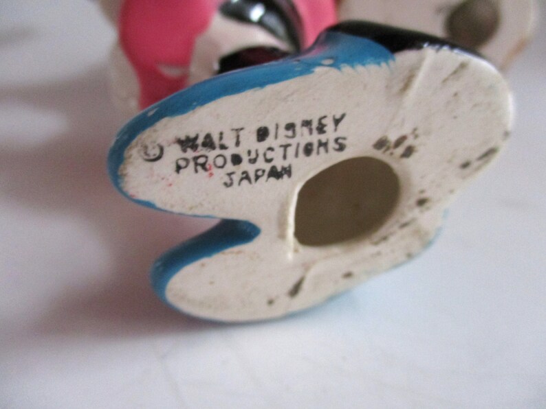 May include: Close-up of the bottom of a ceramic figurine. The figurine is painted blue, black, and pink. The bottom of the figurine is white with black writing that reads "Walt Disney Productions Japan".