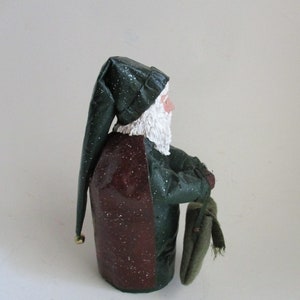 May include: A green and burgundy Santa Claus figurine with a white beard and a green hat. He is holding a green sack in his right hand.
