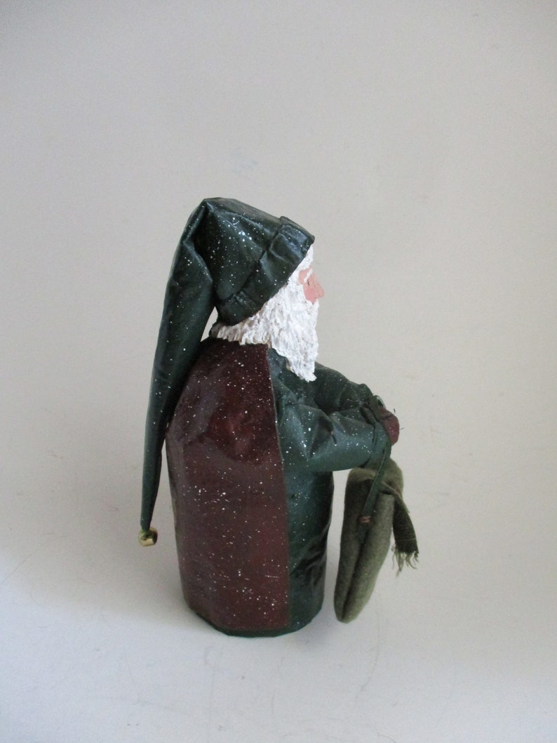 May include: A green and burgundy Santa Claus figurine with a white beard and a green hat. He is holding a green sack in his right hand.