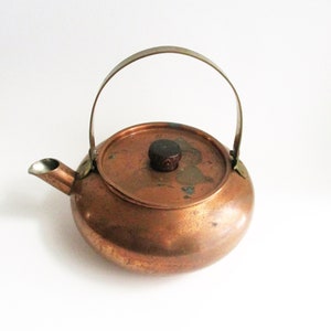 May include: A round copper teapot featuring a curved metal handle, a short spout, and a circular lid topped with a small, dark brown wooden knob. The copper surface displays natural patina and oxidation marks across the body and lid.