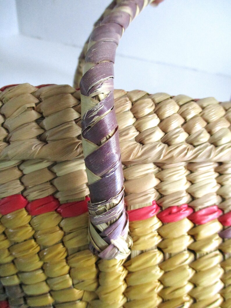 K&ouml;nnte beinhalten: Ein geflochtener Korb mit einem violetten Griff. Der Korb ist aus nat&uuml;rlichen Materialien gefertigt und hat ein rot-gelbes Streifenmuster.