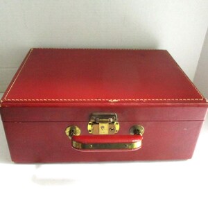 May include: A rectangular red storage case featuring a smooth exterior surface with visible white stitching along the top edges. The case includes a central gold-toned metal latch mechanism and a matching red handle with gold-toned hardware.