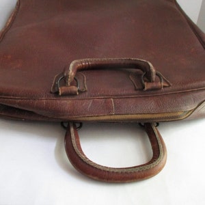 May include: A brown leather briefcase with a top handle and a curved handle on the front. The briefcase has a zipper closure and shows signs of wear.