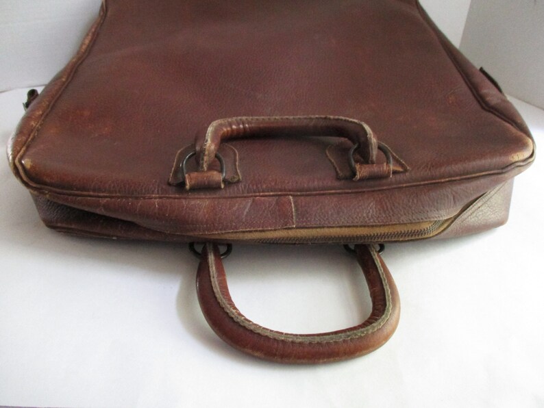 May include: A brown leather briefcase with a top handle and a curved handle on the front. The briefcase has a zipper closure and shows signs of wear.