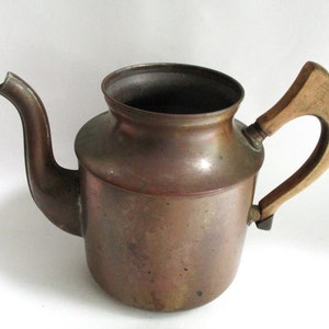 May include: A copper teapot with a weathered, dark metallic finish and a curved spout. The vessel features a wide, open top and a light-colored wooden handle attached to the side with metal hardware, showing signs of age and natural oxidation.