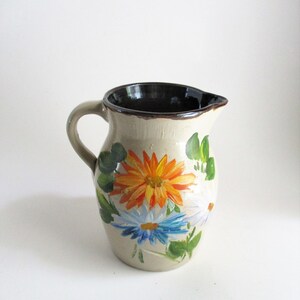 May include: A beige ceramic pitcher with a dark brown interior and handle. The pitcher is decorated with hand-painted flowers in orange, blue, and white, with green leaves. The pitcher has a classic shape and a spout for pouring.