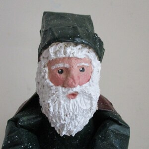 May include: A handmade Santa Claus figurine with a white beard and a green hat with white speckles. The figurine is made of paper mache and has a painted face.