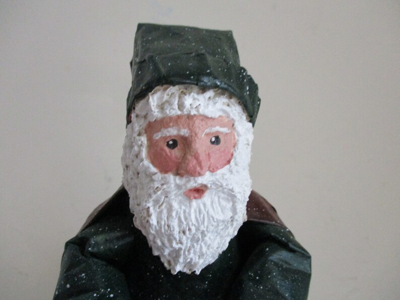 May include: A handmade Santa Claus figurine with a white beard and a green hat with white speckles. The figurine is made of paper mache and has a painted face.