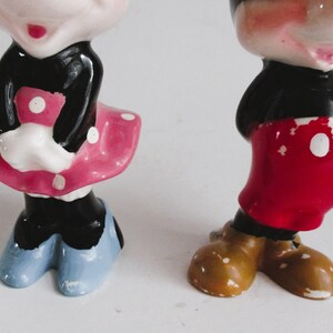 May include: A vintage ceramic figurine set of Mickey and Minnie Mouse. Mickey is wearing red shorts with white polka dots and brown shoes. Minnie is wearing a black dress with a pink skirt and white polka dots and blue shoes.