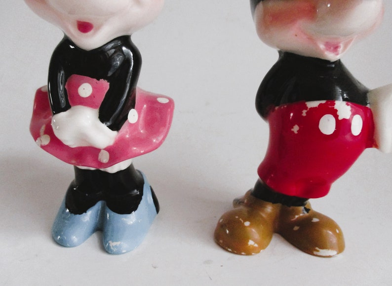 May include: A vintage ceramic figurine set of Mickey and Minnie Mouse. Mickey is wearing red shorts with white polka dots and brown shoes. Minnie is wearing a black dress with a pink skirt and white polka dots and blue shoes.