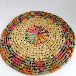 May include: A round, woven placemat with a natural beige center and a colorful border. The border features hues of orange, pink, green, and purple. The center has a starburst design in the center. This decorative item is perfect for adding a touch of rustic charm to any table setting.