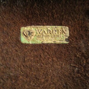 May include: Close-up of a brown, textured surface with a rectangular, faded green and white label. The label reads "WARREN LEATHER GOODS CO WORCESTER MASS".