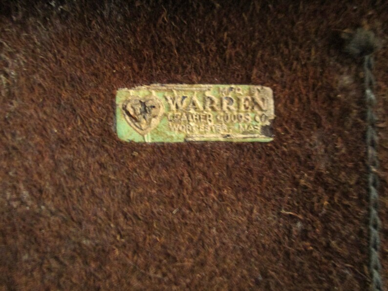 May include: Close-up of a brown, textured surface with a rectangular, faded green and white label. The label reads "WARREN LEATHER GOODS CO WORCESTER MASS".