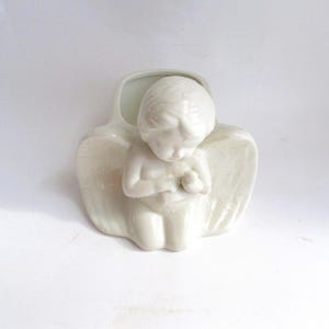 May include: A white ceramic angel figurine with folded wings and a serene expression. The angel is kneeling, with hands clasped in prayer. The figurine has a smooth, glossy finish and is set against a white background.