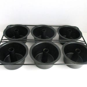 May include: A black metal baking rack with six individual angel food cake pans. Each pan has a central tube and a rounded base. The rack is designed to hold the pans securely.
