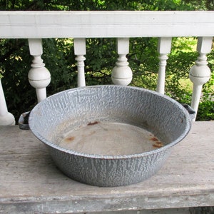 May include: A vintage, round, gray enamelware basin with a textured surface and two small handles. The basin sits on a weathered wooden surface, with a white railing and green foliage in the background. This antique piece is ideal for farmhouse decor.