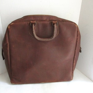 May include: A brown leather briefcase with a top handle and two smaller handles. The bag has a rectangular shape with rounded corners and a zipper closure. The leather has a worn appearance.