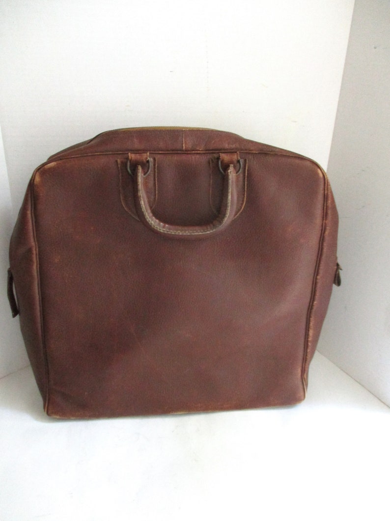 May include: A brown leather briefcase with a top handle and two smaller handles. The bag has a rectangular shape with rounded corners and a zipper closure. The leather has a worn appearance.