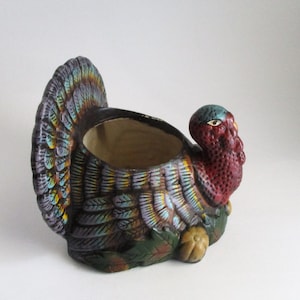 May include: A ceramic turkey-shaped planter featuring a dark brown body with iridescent blue and yellow highlights on the tail feathers. The turkey has a textured red head and neck, and sits atop a base decorated with green leaves and a small yellow gourd.