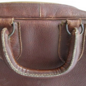 May include: Close-up of a brown leather bag with a handle. The bag has a zipper closure and metal rings attaching the handle. The leather shows signs of wear, suggesting it is vintage or well-used.