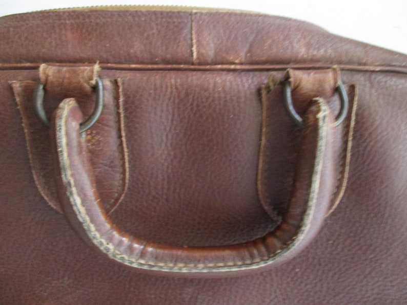 May include: Close-up of a brown leather bag with a handle. The bag has a zipper closure and metal rings attaching the handle. The leather shows signs of wear, suggesting it is vintage or well-used.