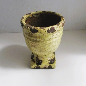 May include: A yellow ceramic planter with a crackled glaze and brown accents. The planter has a wide, round top and a square base.