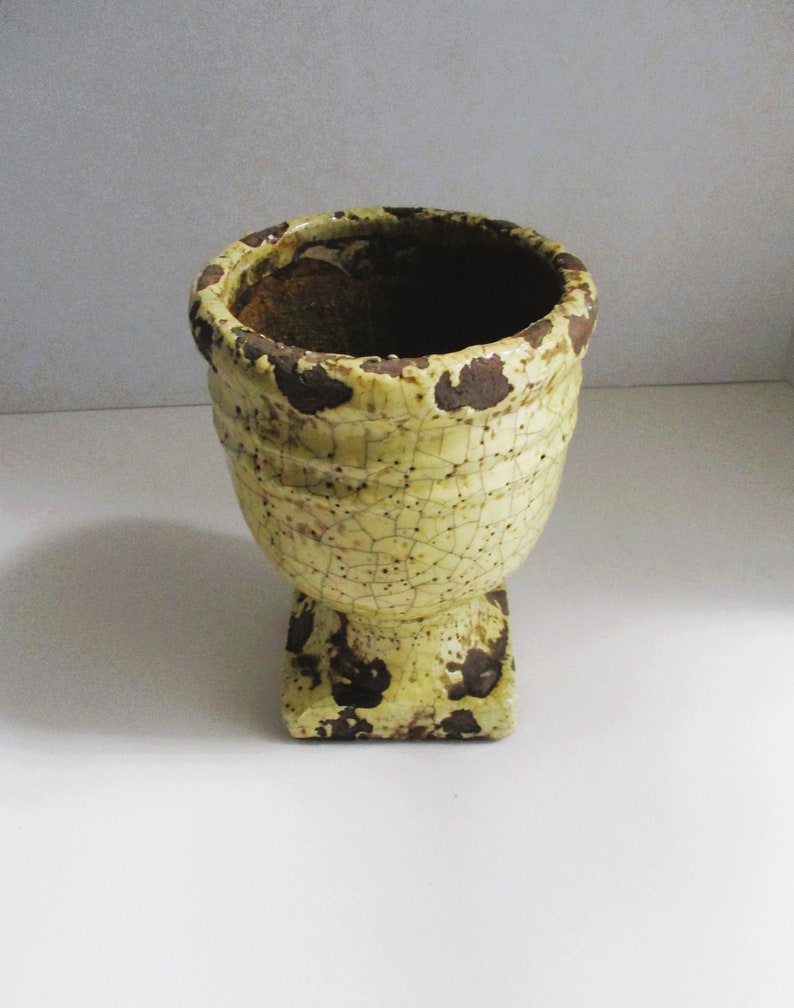 May include: A yellow ceramic planter with a crackled glaze and brown accents. The planter has a wide, round top and a square base.