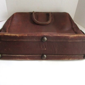 May include: A vintage brown leather briefcase with a top handle and metal feet. The bag has a rectangular shape with rounded edges and shows signs of wear, adding to its antique charm. The handle is a darker shade of brown.