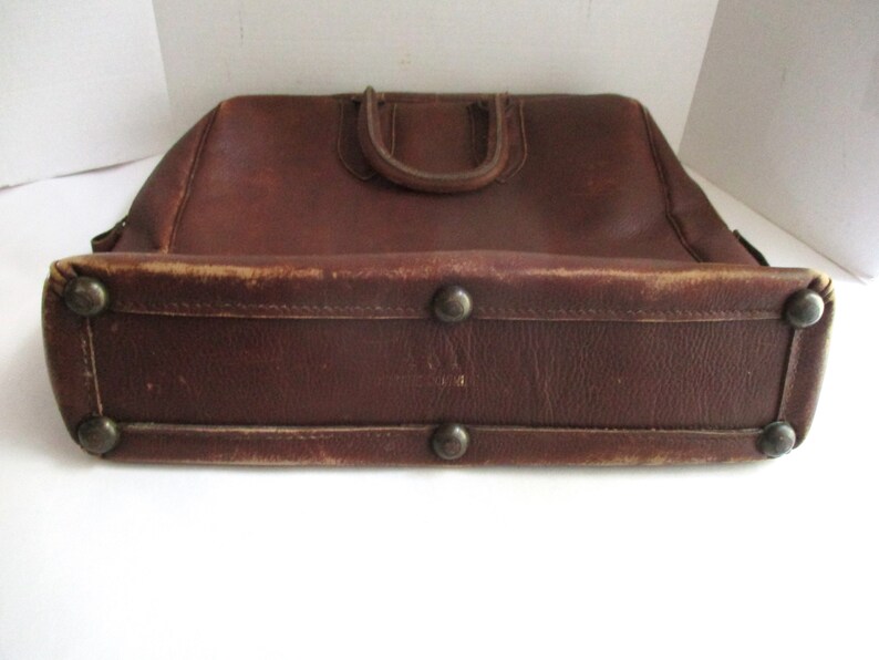 May include: A vintage brown leather briefcase with a top handle and metal feet. The bag has a rectangular shape with rounded edges and shows signs of wear, adding to its antique charm. The handle is a darker shade of brown.