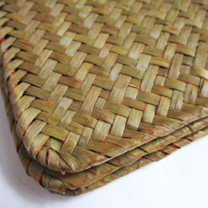 May include: Close-up of a woven mat or basket with a repeating pattern. The natural fibers are a mix of light and medium brown, with red accents visible along the edges. The texture is tightly woven and appears durable.