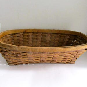 May include: A woven oval basket with a light brown color. The basket is made of woven strips of wood, with a sturdy rim. The basket is empty and sits on a white surface. The basket is likely used for storage or display.