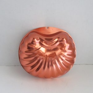 May include: A copper-colored, vintage bundt cake mold with a scalloped edge and a central design. The mold has a hole at the top for hanging. The shiny surface reflects light, highlighting the intricate details of the design.