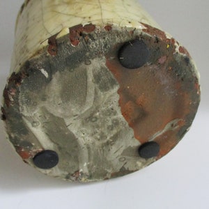 May include: The base of a ceramic vase with a white glaze and brown clay showing through. There are three black rubber feet on the bottom.