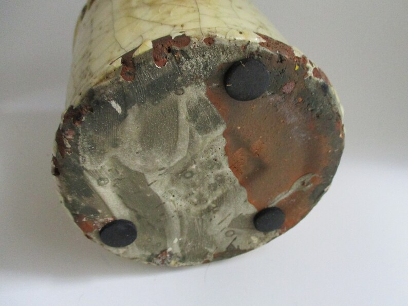 May include: The base of a ceramic vase with a white glaze and brown clay showing through. There are three black rubber feet on the bottom.