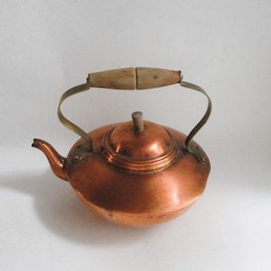 May include: A copper teapot with a rounded body, a spout, and a wooden handle. The handle is attached to the pot with brass brackets. The lid is also copper and has a small wooden knob. The teapot is designed for stovetop use.