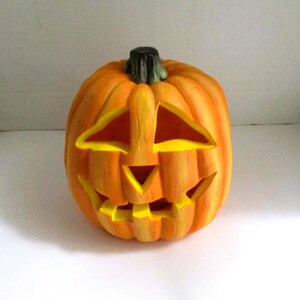 May include: A carved orange pumpkin jack-o'-lantern with a dark green stem. The pumpkin features triangular eyes, a triangular nose, and a wide, jagged mouth with visible teeth. The interior of the carved openings is painted a bright yellow.