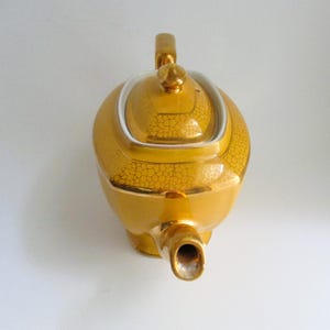 May include: A gold teapot with a textured, crackled design. The teapot has a rounded body, a curved handle, and a spout. The lid is topped with a small finial. The teapot is likely made of ceramic or porcelain.