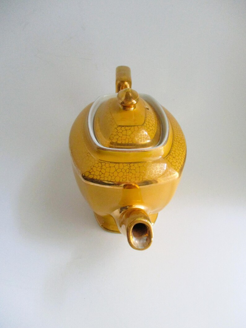May include: A gold teapot with a textured, crackled design. The teapot has a rounded body, a curved handle, and a spout. The lid is topped with a small finial. The teapot is likely made of ceramic or porcelain.