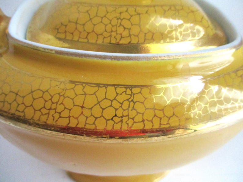 May include: Close-up of a yellow ceramic teapot with a lid. The teapot has a gold, cracked pattern design and a white rim. The teapot is a light yellow colour and has a rounded shape.